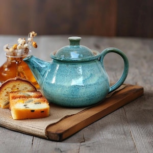 Puede incluir: Una tetera de cerámica turquesa con tapa y asa a juego descansa sobre una tabla de madera. Rebanadas de pan y un tarro de miel están en el fondo. La tetera tiene un esmalte moteado.