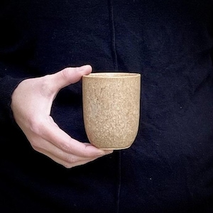 May include: A beige ceramic mug with a speckled texture. The mug is held in a hand.