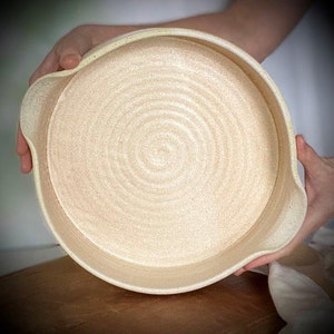 Puede incluir: Un plato de servir redondo de cerámica blanca con un borde ligeramente elevado. El plato tiene una superficie texturizada con un sutil patrón de remolino.