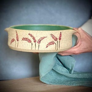 Puede incluir: Un plato para hornear redondo de cerámica con un esmalte beige claro y un esmalte verde en el interior. El plato tiene dos asas y está decorado con espigas de trigo rojas.
