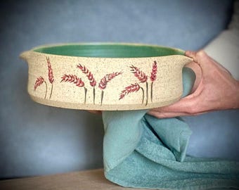 Può includere: Una teglia da forno rotonda in ceramica con una glassa beige chiaro e una glassa verde all'interno. La teglia ha due manici ed è decorata con spighe di grano rosse.
