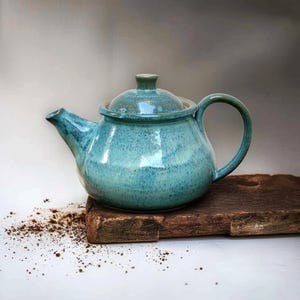 Puede incluir: Una tetera de cerámica turquesa con tapa y asa a juego. La tetera tiene un cuerpo redondeado y un pico para verter. El esmalte tiene un aspecto moteado. La tetera está sobre una superficie de madera.