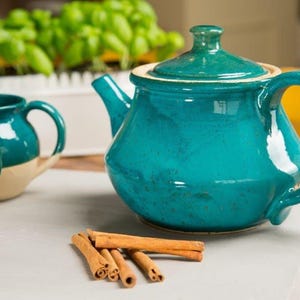May include: A teal blue ceramic teapot with a lid and a matching set of two mugs. The teapot and mugs have a white rim and are sitting on a white surface with cinnamon sticks.