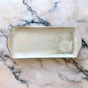 Puede incluir: Una bandeja rectangular de cerámica con bordes redondeados. La bandeja tiene una superficie moteada de color blanquecino con sutiles tonos verdes y marrones. El borde es de color marrón oscuro. Adecuada para servir aperitivos o platos pequeños.