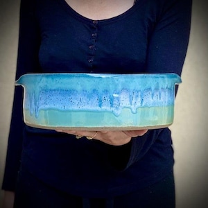 Puede incluir: Un plato para hornear de cerámica grande, de forma ovalada, con un glaseado azul claro. El glaseado tiene un efecto moteado, con tonos de azul, verde y blanco. El plato tiene un borde ligeramente elevado.