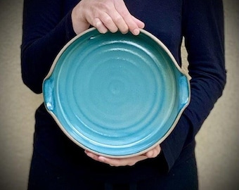 May include: A large, teal blue ceramic serving dish with two handles. The dish has a slightly raised rim and a smooth, glazed finish.