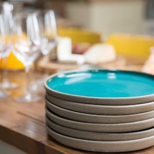 Puede incluir: Una pila de seis platos de cerámica con un centro azul turquesa y un borde gris. Los platos están sobre una superficie de madera, con copas de vino y alimentos en el fondo. Ideal para servir aperitivos o una comida.