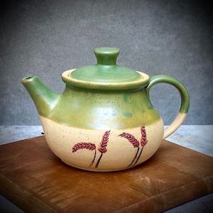 May include: A green and beige ceramic teapot with a lid and a handle. The teapot has a speckled glaze and a design of red wheat stalks on the side.
