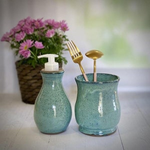 May include: A set of two teal ceramic kitchen containers. One container is a soap dispenser with a white pump. The other container is a utensil holder with a gold fork and spoon inside.