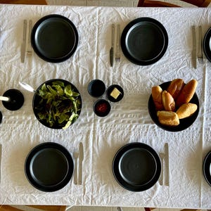 Puede incluir: Vista aérea de una mesa de comedor preparada para una comida. La mesa está cubierta con un mantel blanco y está puesta con platos negros, cubiertos y cuencos de comida, incluyendo ensalada y pan. También hay candelabros negros.