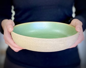 May include: A large, oval ceramic serving bowl with a light green glaze on the inside and a speckled beige glaze on the outside. The bowl has a gold rim.
