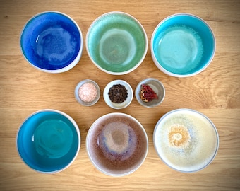 May include: A collection of ceramic bowls in various colors, including blue, green, and brown, arranged on a wooden surface. Small bowls contain salt, pepper, and dried tomatoes. The bowls have a glossy finish and white rims.