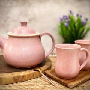 May include: A pink teapot and two mugs with a speckled glaze. The teapot has a rounded body and a curved handle. The mugs have a similar shape and are slightly taller than the teapot. The set is on a wooden tray with a gold spoon.