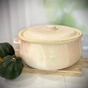 May include: A ceramic casserole dish with a lid, in shades of cream and peach, sits on a wooden cutting board. Two small green and orange gourds are next to the dish. The dish has two handles and a knob on the lid.