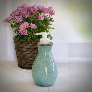 May include: A teal ceramic soap dispenser with a white pump. The dispenser is sitting on a white surface in front of a potted plant with pink flowers.