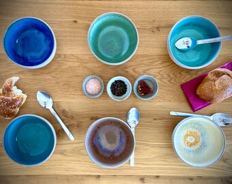 May include: A set of six ceramic bowls in various shades of blue, green, and brown. The bowls are arranged on a wooden surface with small bowls of salt, peppercorns, and red pepper flakes. A pretzel and a roll are also on the surface.