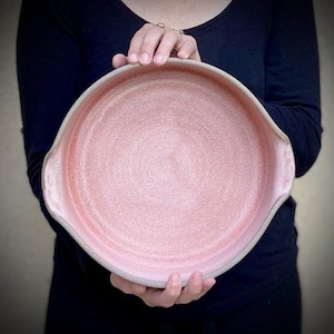 May include: A large, round, pink ceramic baking dish with two handles. The dish has a speckled glaze and a white rim.