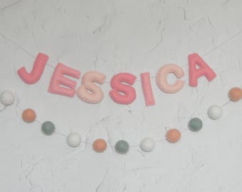 May include: A pink felt banner with the name "JESSICA" spelled out in pink felt letters. The banner is strung with a white string and decorated with small white, peach, and green felt balls.