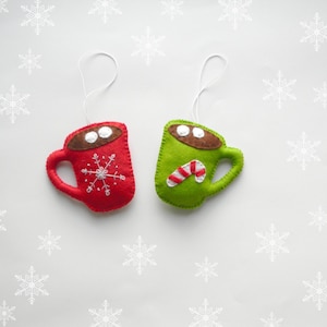 May include: Two felt Christmas decorations shaped like mugs. One mug is red with a snowflake design and the other is green with a candy cane design.