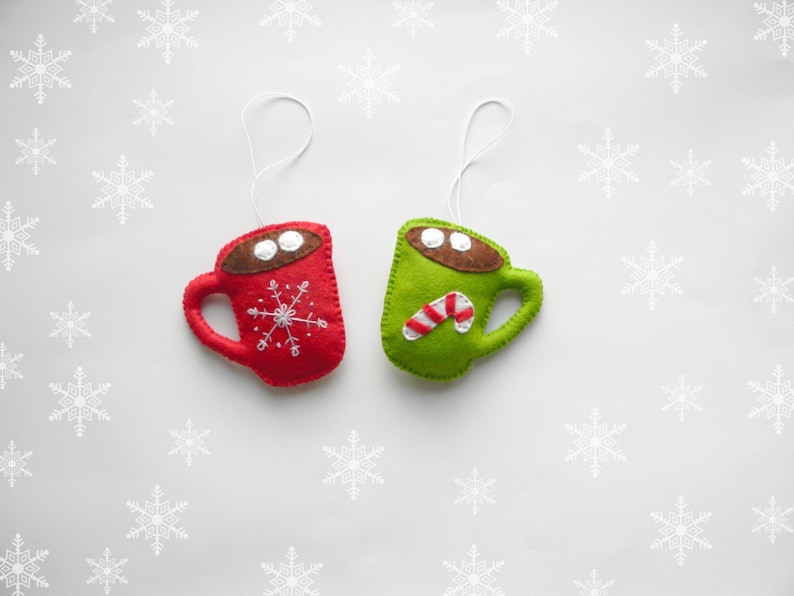 May include: Two felt Christmas decorations shaped like mugs. One mug is red with a snowflake design and the other is green with a candy cane design.