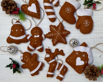 May include: A collection of brown felt Christmas ornaments, including a gingerbread man, heart, candy cane, snowflake, bell, stocking, mitten, house, and reindeer. Each ornament has white accents and is strung with twine. Silver bells and pinecones are also present.