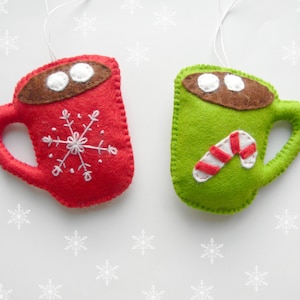 May include: Two felt Christmas decorations shaped like mugs. One mug is red with a white snowflake and the other is green with a white candy cane.