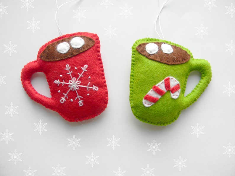 May include: Two felt Christmas decorations shaped like mugs. One mug is red with a white snowflake and the other is green with a white candy cane.