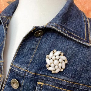 May include: A white and gold brooch with a floral design is pinned to a dark blue denim jacket. The brooch features white, oval-shaped stones and small, clear crystals. The jacket has a classic collar, button closures, and visible stitching.