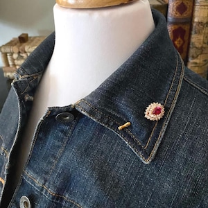 May include: A denim jacket collar with a ruby and diamond brooch. The brooch features a central red gemstone surrounded by small, clear stones. The jacket is a dark blue wash, and the brooch is pinned to the collar.