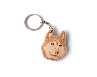 May include: A wooden keychain featuring a detailed engraving of a Husky dog's face. The keychain has a silver-colored metal ring and chain. The dog's fur is depicted with intricate lines, and the eyes and nose are clearly defined.