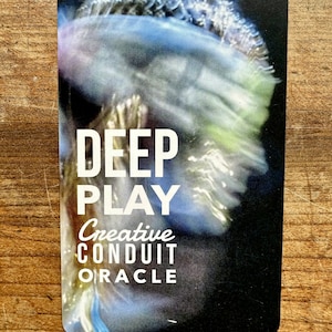 Könnte beinhalten: Eine Karte mit einem verschwommenen Bild eines blauen und grünen Lichtmusters. Der Text "DEEP PLAY" ist in weißen Buchstaben auf schwarzem Hintergrund. Unter dem Text steht "Creative Conduit Oracle" in weißen Buchstaben.