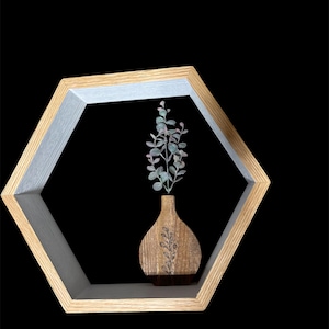 Peut inclure: Une étagère hexagonale en bois avec un cadre en bois clair et un intérieur blanc. Un petit vase en bois avec un motif de feuilles contient une branche de verdure. L'étagère est sur fond noir.