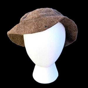 May include: A brown, textured hat with a small brim, displayed on a white mannequin head against a black background. The hat appears to be made of a woven material, possibly wool or a similar fabric. The hat's colour is a warm, earthy brown.