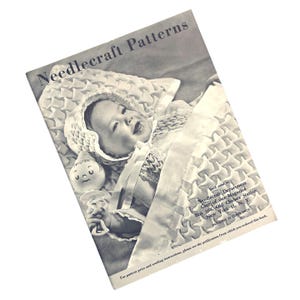 May include: A vintage Needlecraft Patterns booklet featuring a black and white photograph of an infant wearing a smocked bonnet and holding a small toy rattle. The cover includes text with mailing instructions for the Needlecraft Department.