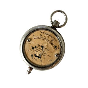 May include: An antique silver-toned ammeter with a circular face. The face has a paper background with black markings and the text "EVER READY" at the top. The gauge reads "AMPERES" and "AMERICAN ELEC. NOV. & MFG. CO. NEW YORK." A metal loop is at the top.