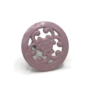 May include: A pink metal cabinet knob with a decorative, openwork design. The knob is round and has a distressed finish.