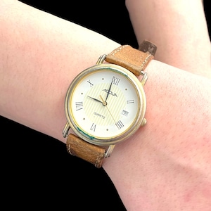 Puede incluir: Un reloj de pulsera dorado con esfera blanca y números romanos. El reloj tiene una correa de cuero marrón y la palabra "ACQUA" en la esfera. El reloj también tiene una pantalla de fecha. El reloj es de cuarzo.