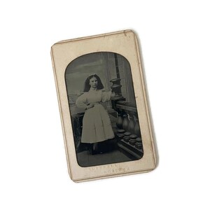 May include: Antique black and white photograph of a young person in a long dress, standing on a balcony. The photograph is in a rectangular frame with an arched top. The text at the bottom of the image is blurry.