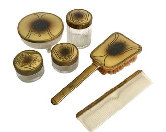 May include: A vintage vanity set with a gold-tone finish. The set includes a round powder dish, three jars, a brush with a gold handle, and a comb.