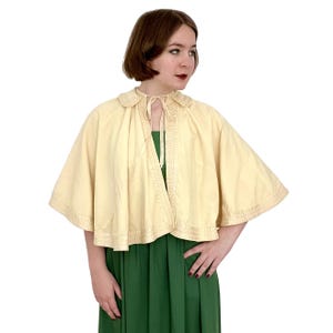 May include: A cream-colored cape with a satin trim and a satin ribbon tie. The cape has a collar and is made of a soft, lightweight fabric.