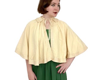 May include: A cream-colored cape with a satin trim and a satin ribbon tie. The cape has a collar and is made of a soft, lightweight fabric.