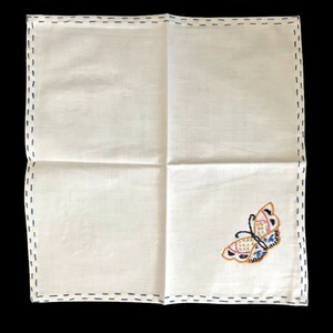 May include: White linen handkerchief with a blue stitched border. The handkerchief features an embroidered butterfly in shades of pink, blue, and gold. The butterfly is positioned in the lower right corner.
