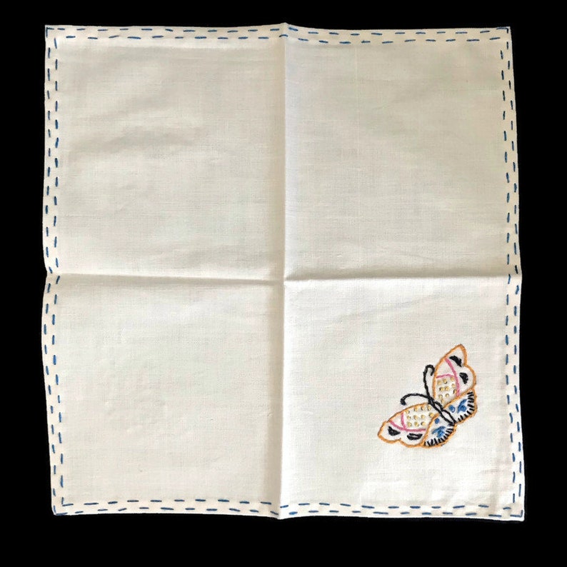 May include: White linen handkerchief with a blue stitched border. The handkerchief features an embroidered butterfly in shades of pink, blue, and gold. The butterfly is positioned in the lower right corner.