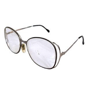 Może przedstawiać: Okulary w stylu vintage z metalową, srebrną oprawą i dużymi, okrągłymi soczewkami. Okulary mają podwójną obręcz wokół soczewek i czarne zauszniki. Noski są w jasnobeżowym kolorze. Okulary są na białym tle.