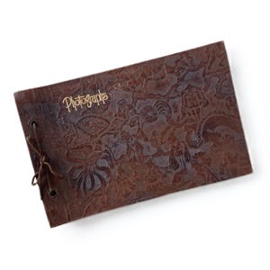 Puede incluir: Un álbum de fotos de cuero marrón con un diseño en relieve y la palabra "Fotografías" en letras doradas en la portada.