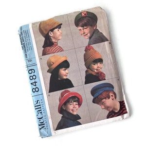 May include: A vintage sewing pattern for children's hats. The pattern includes five different hat styles: a newsboy cap, a beanie with a pom-pom, a cloche hat, a bucket hat, and a sailor cap. The pattern is printed on a white background with black text and illustrations. The pattern is labelled "McCall's 8489" and "Hat Wardrobe for Children, Boys and Girls".