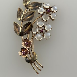 May include: A gold tone brooch with a floral design. The brooch features three flowers with moonstone cabochons and ruby accents. The flowers are set on a stem with leaves.