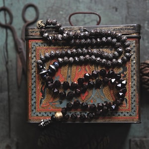 May include: A vintage tin box with a colorful floral design, topped with a black and gray beaded necklace. The necklace has a gold charm and a silver pendant.