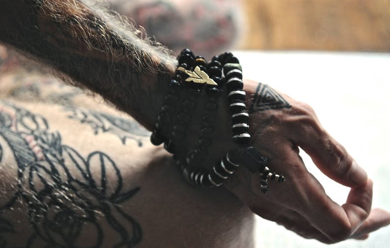 K&ouml;nnte beinhalten: Eine Person tr&auml;gt einen Stapel schwarzer Perlenarmb&auml;nder mit einem goldenen Akzent am Handgelenk. Die Person hat T&auml;towierungen am Arm und an der Hand.