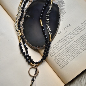 May include: A black and gold beaded necklace with a circular pendant. The necklace is lying on an open book with the text "LOCRINE" visible.